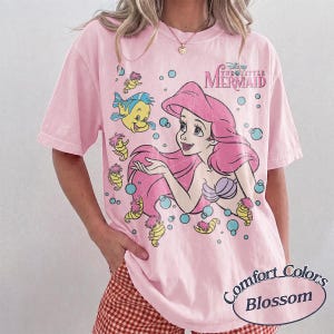 May include: A light pink short-sleeve t-shirt featuring a graphic print of Ariel from The Little Mermaid with Flounder and seahorses. The design includes blue bubbles and the text The Little Mermaid in pink. The shirt has a crew neckline.