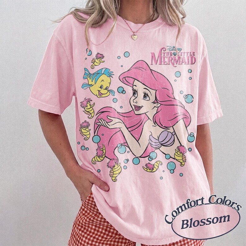 May include: A light pink short-sleeve t-shirt featuring a graphic print of Ariel from The Little Mermaid with Flounder and seahorses. The design includes blue bubbles and the text The Little Mermaid in pink. The shirt has a crew neckline.