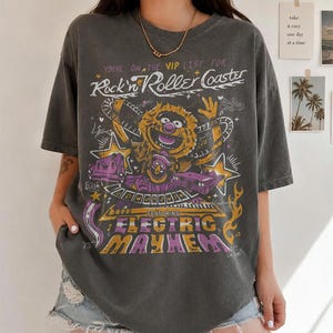 Puede incluir: Camiseta gris oscuro con un dise&ntilde;o gr&aacute;fico que presenta un personaje de los Muppets y el texto "Rock'n Roller Coaster" y "Electric Mayhem". El dise&ntilde;o incorpora elementos morados, amarillos y blancos. La camiseta tiene mangas cortas y cuello redondo.