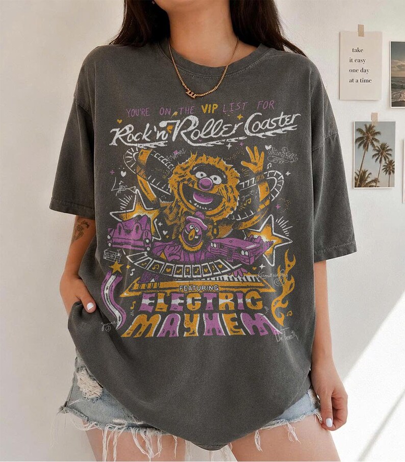 Puede incluir: Camiseta gris oscuro con un dise&ntilde;o gr&aacute;fico que presenta un personaje de los Muppets y el texto "Rock'n Roller Coaster" y "Electric Mayhem". El dise&ntilde;o incorpora elementos morados, amarillos y blancos. La camiseta tiene mangas cortas y cuello redondo.