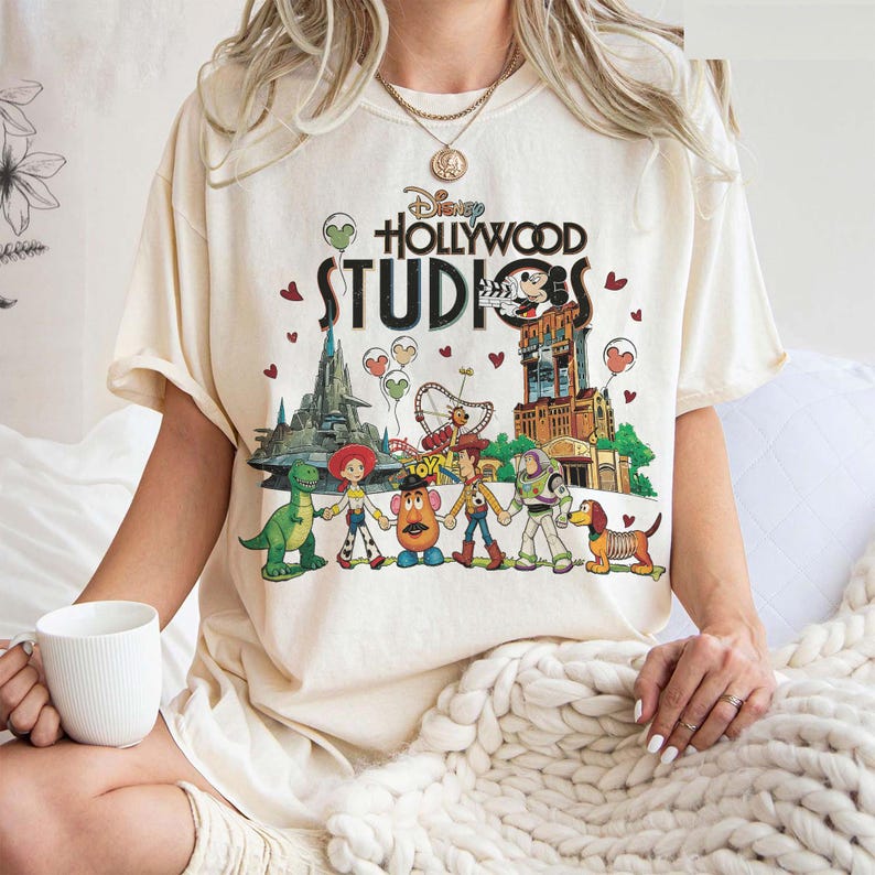May include: Cream-colored t-shirt featuring the Disney Hollywood Studios logo and a colorful graphic of Disney characters. The graphic includes characters from Toy Story, a dinosaur, and a building with Mickey Mouse.
