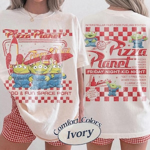 K&ouml;nnte beinhalten: Cremefarbenes T-Shirt mit einem Pizza Planet-Design, das drei gr&uuml;ne Aliens in blauen Overalls zeigt. Auf der Vorderseite steht "FOOD & FUN SPACE PORT". Auf der R&uuml;ckseite steht "INTERSTELLAR FAST-FOOD FUELING STATION."