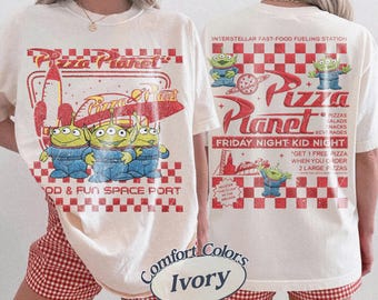 May include: Ivory-colored t-shirt featuring a Pizza Planet graphic with three green aliens in blue overalls. The front design includes the text "FOOD & FUN SPACE PORT." The back of the shirt has the text "INTERSTELLAR FAST-FOOD FUELING STATION."