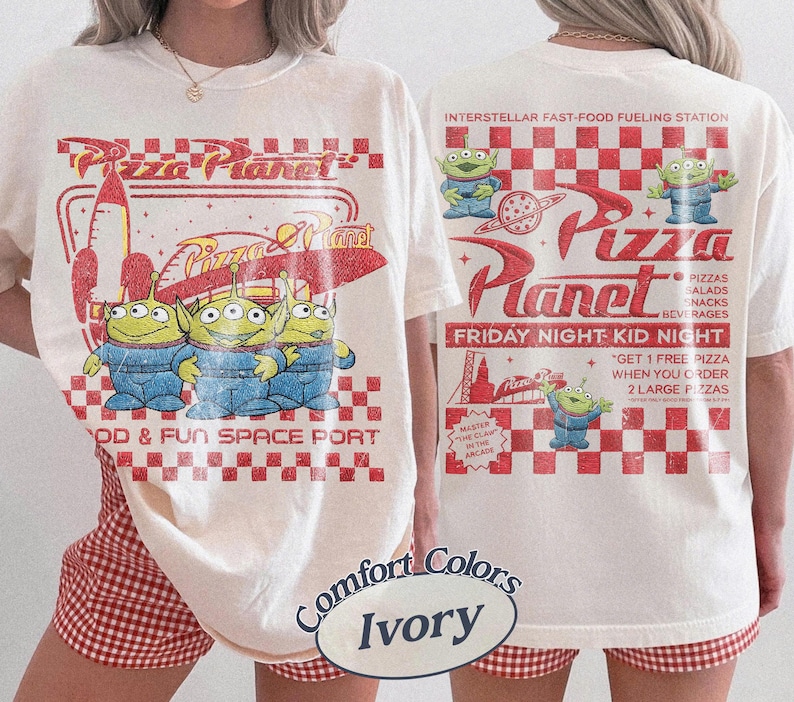 K&ouml;nnte beinhalten: Cremefarbenes T-Shirt mit einem Pizza Planet-Design, das drei gr&uuml;ne Aliens in blauen Overalls zeigt. Auf der Vorderseite steht "FOOD & FUN SPACE PORT". Auf der R&uuml;ckseite steht "INTERSTELLAR FAST-FOOD FUELING STATION."