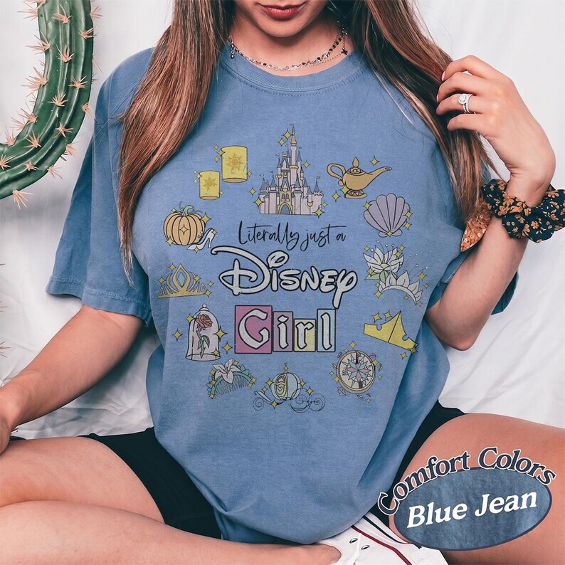 Op de afbeelding: Een blauwe jeanskleurige t-shirt met de tekst "Literally just a Disney Girl" en diverse Disney-thema graphics. De graphics omvatten een kasteel, een lamp, een pompoen en een zeeschelp. Het shirt heeft ook de woorden "Comfort Colors" en "Blue Jean" erop gedrukt.