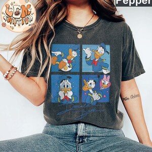 May include: A dark grey t-shirt with a vintage cartoon print of Donald Duck in four different poses. The shirt has a distressed look and the print is in blue, yellow, and red. The text "Donald Duck" is printed below the cartoon.