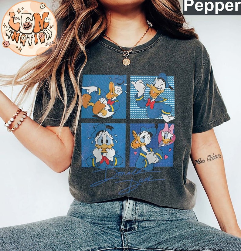 May include: A dark grey t-shirt with a vintage cartoon print of Donald Duck in four different poses. The shirt has a distressed look and the print is in blue, yellow, and red. The text "Donald Duck" is printed below the cartoon.