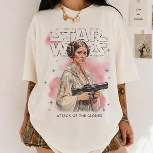 K&ouml;nnte beinhalten: Cremefarbenes T-Shirt mit einer Grafik von Prinzessin Leia aus Star Wars, die einen Blaster h&auml;lt, mit dem Text "STAR WARS" und "ATTACK OF THE CLONES". Das Design enth&auml;lt rosa Akzente und Sterne.