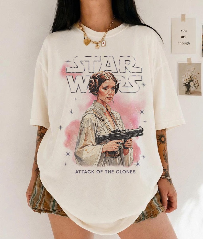 K&ouml;nnte beinhalten: Cremefarbenes T-Shirt mit einer Grafik von Prinzessin Leia aus Star Wars, die einen Blaster h&auml;lt, mit dem Text "STAR WARS" und "ATTACK OF THE CLONES". Das Design enth&auml;lt rosa Akzente und Sterne.