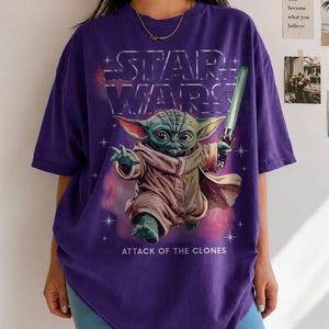 K&ouml;nnte beinhalten: Lila Star Wars T-Shirt mit Yoda, der ein gr&uuml;nes Lichtschwert h&auml;lt. Das silberne Star Wars Logo und der Text "ATTACK OF THE CLONES" sind ebenfalls zu sehen. Das T-Shirt hat kurze &Auml;rmel und eine lockere Passform.
