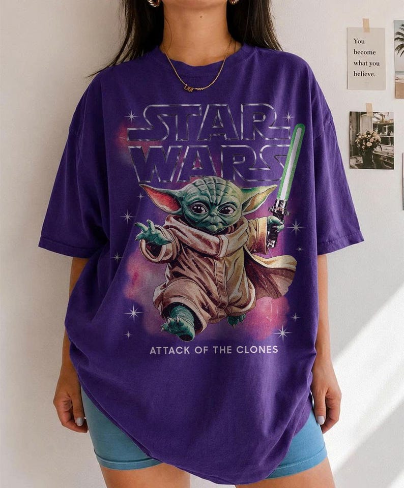 K&ouml;nnte beinhalten: Lila Star Wars T-Shirt mit Yoda, der ein gr&uuml;nes Lichtschwert h&auml;lt. Das silberne Star Wars Logo und der Text "ATTACK OF THE CLONES" sind ebenfalls zu sehen. Das T-Shirt hat kurze &Auml;rmel und eine lockere Passform.