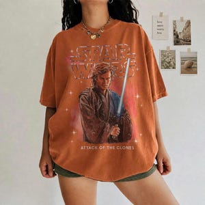 K&ouml;nnte beinhalten: Gebrannt orangefarbenes Star Wars T-Shirt mit Anakin Skywalker und einem Lichtschwert. Das Shirt hat das Star Wars Logo und den Text "ATTACK OF THE CLONES". Das Shirt ist kurz&auml;rmlig und oversized.