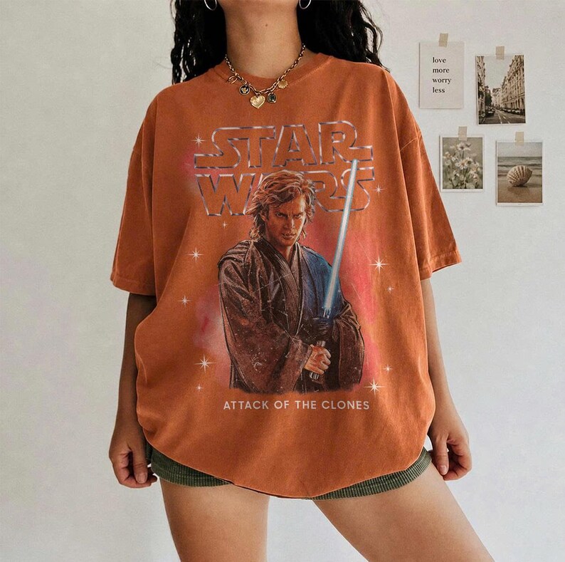 K&ouml;nnte beinhalten: Gebrannt orangefarbenes Star Wars T-Shirt mit Anakin Skywalker und einem Lichtschwert. Das Shirt hat das Star Wars Logo und den Text "ATTACK OF THE CLONES". Das Shirt ist kurz&auml;rmlig und oversized.