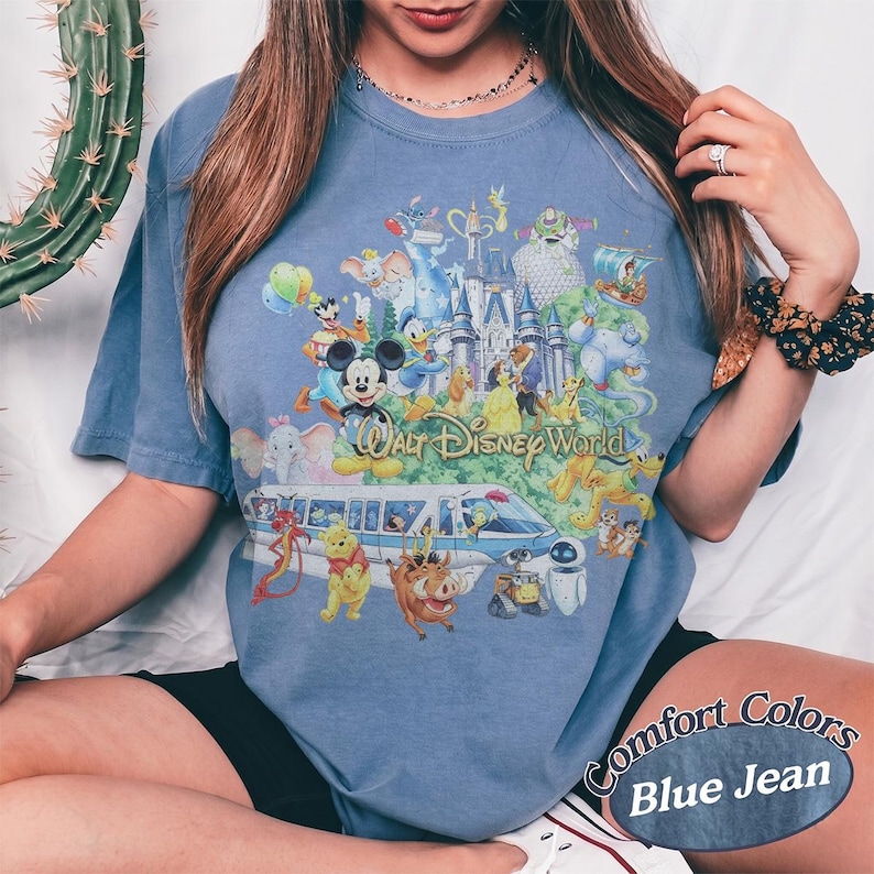 May include: A blue jean colored t-shirt featuring a large graphic print of Walt Disney World with various animated characters, a castle, and a monorail. The text Walt Disney World is printed in the center of the colorful character collage.