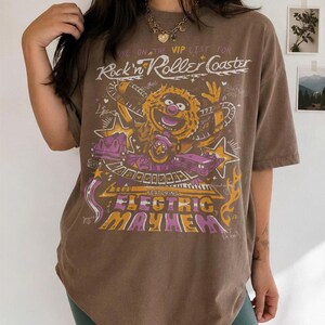 Puede incluir: Camiseta marr&oacute;n con un gr&aacute;fico de un personaje de los Muppets y el texto "Rock'n Roller Coaster" y "Electric Mayhem". La camiseta tiene un aspecto vintage.
