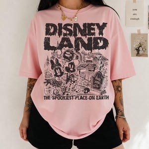 May include: Pale pink t-shirt featuring a black graphic with the words "DISNEY LAND" and a spooky illustration of Mickey and Minnie Mouse. The text "THE SPOOKIEST PLACE ON EARTH" is below the graphic.