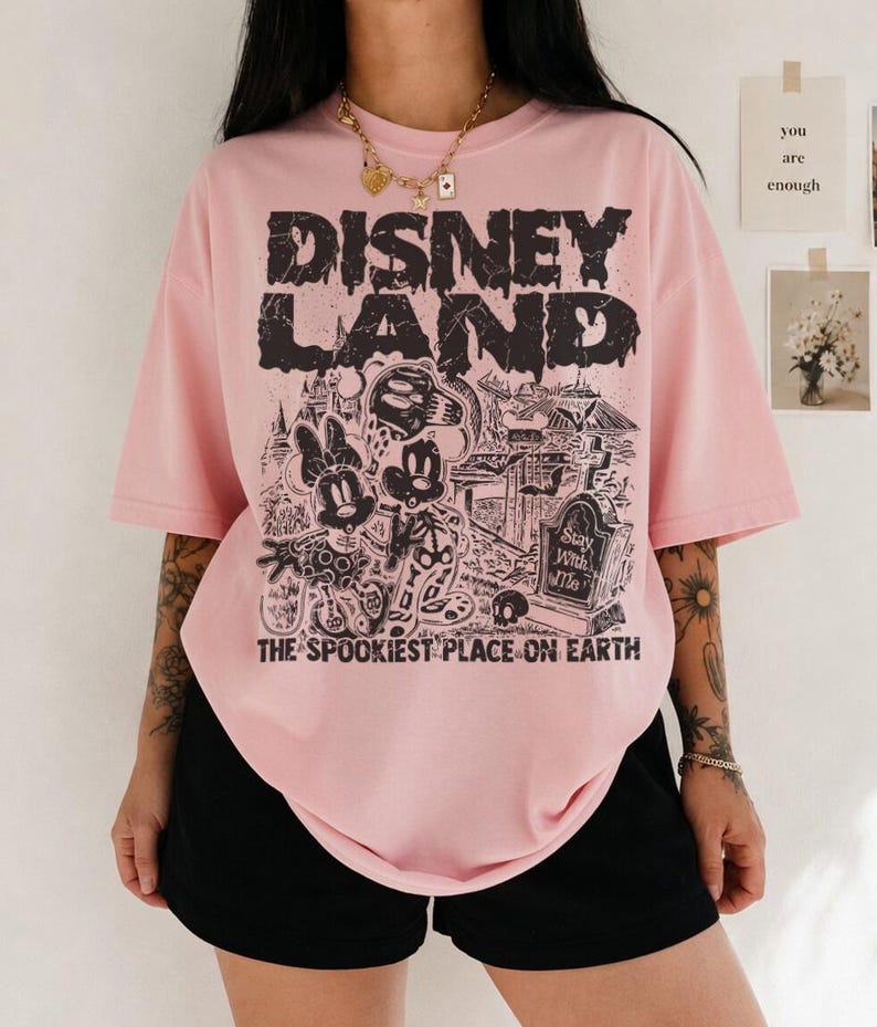 May include: Pale pink t-shirt featuring a black graphic with the words "DISNEY LAND" and a spooky illustration of Mickey and Minnie Mouse. The text "THE SPOOKIEST PLACE ON EARTH" is below the graphic.