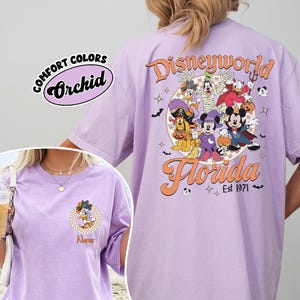 May include: A light purple t-shirt with a Disney World Florida graphic. The back features Disney characters in Halloween costumes. The front has a smaller graphic of a character and the word "Name". The shirt is labeled "Comfort Colors Orchid".