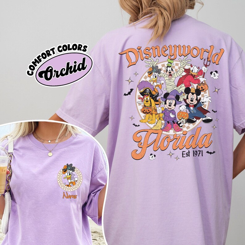 May include: A light purple t-shirt with a Disney World Florida graphic. The back features Disney characters in Halloween costumes. The front has a smaller graphic of a character and the word "Name". The shirt is labeled "Comfort Colors Orchid".