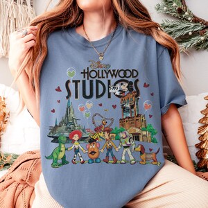 Op de afbeelding: Lichtblauw T-shirt met de tekst "Disney Hollywood Studios" en een afbeelding van Disney-personages en parkattracties. De afbeelding bevat Mickey Mouse, Toy Story-personages en andere parkelementen.