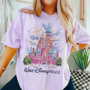May include: A light purple t-shirt featuring a colorful graphic of Cinderella's castle at Walt Disney World. The graphic includes the text "This is Now!" and "Walt Disney World 1997". The castle is pink with blue and red accents.