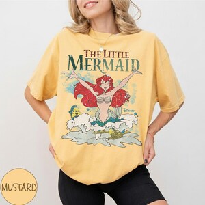 May include: A mustard yellow short-sleeve t-shirt featuring a graphic print of The Little Mermaid with Ariel, Flounder, and Sebastian. The text reads The Little Mermaid in a dark green font. The design includes water splashes and bubbles.