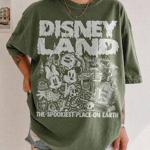 May include: An olive green t-shirt featuring the text "DISNEY LAND" in a distressed white font, with a spooky illustration of Mickey and Minnie Mouse. The text "THE SPOOKIEST PLACE ON EARTH" is below the graphic.