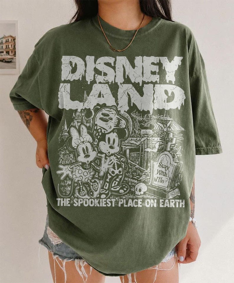 May include: An olive green t-shirt featuring the text "DISNEY LAND" in a distressed white font, with a spooky illustration of Mickey and Minnie Mouse. The text "THE SPOOKIEST PLACE ON EARTH" is below the graphic.
