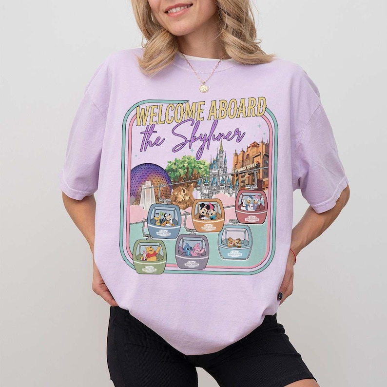 May include: A light purple t-shirt with a graphic featuring the text "WELCOME ABOARD the Skyliner". The graphic includes images of Disney characters in gondolas, with park landmarks in the background.