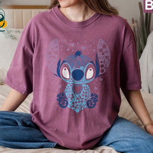 May include: A mauve t-shirt with a blue and purple floral design featuring Stitch from Lilo & Stitch. The design includes hearts and flowers.
