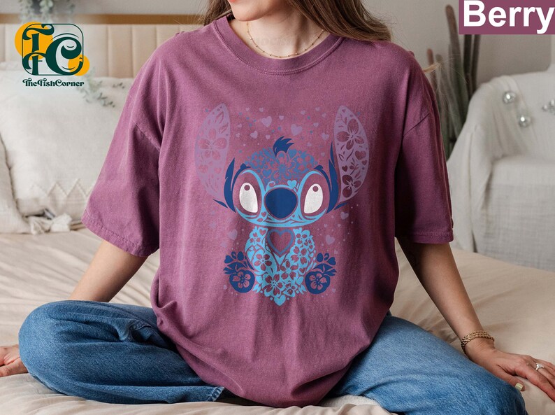May include: A mauve t-shirt with a blue and purple floral design featuring Stitch from Lilo & Stitch. The design includes hearts and flowers.