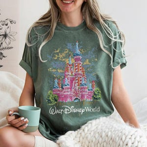 May include: Green t-shirt featuring a pink castle design with blue and white accents, and the text "Walt Disney World". The castle has a striped tower and is surrounded by green bushes. The shirt is a comfortable fit.