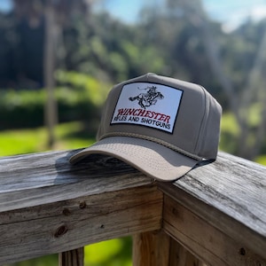 Può includere: Un cappellino da baseball beige con visiera piatta e dettaglio corda. Il cappellino presenta una toppa rettangolare con la scritta "Winchester Rifles and Shotguns" in rosso e bianco, e una grafica di una persona a cavallo.