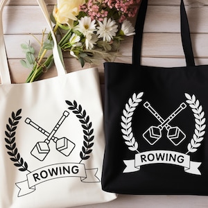 Puede incluir: Dos bolsas de lona blancas con asas negras. Las bolsas tienen un diseño en blanco y negro de remos cruzados con una corona de laurel y la palabra "ROWING" en una pancarta. Una bolsa es blanca con diseño negro y la otra es negra con diseño blanco.