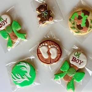 Pode incluir: Variedade de biscoitos de açúcar decorados em sacos de celofane transparentes. Os desenhos incluem um ursinho de pelúcia, uma cabeça de veado, uma pegada e biscoitos com o texto "Oh Boy" e um laço. Os biscoitos são decorados com glacê verde, castanho e branco.