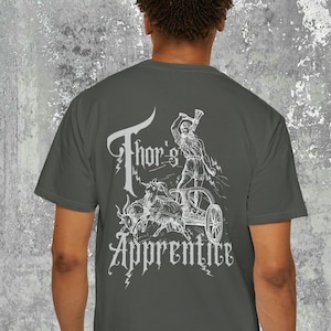 Puede incluir: Una camiseta gris oscuro con un estampado gráfico blanco de un hombre conduciendo un carro tirado por dos cabras. El texto "Thor's Apprentice" está impreso debajo del gráfico.