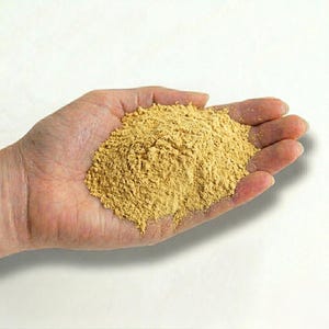 May include: A hand holding a pile of fine, light brown powder. The powder is a uniform color and appears to be a dry, granular substance. The background is plain white.