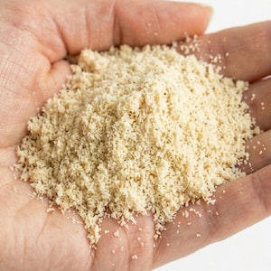 K&ouml;nnte beinhalten: Nahaufnahme einer Hand, die einen Haufen fein gemahlenes, hellbeige Pulver h&auml;lt. Das Pulver hat eine leicht k&ouml;rnige Textur und scheint ein Lebensmittel zu sein, m&ouml;glicherweise Mandelmehl oder eine &auml;hnliche Zutat zum Backen oder Kochen.