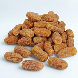 Puede incluir: Un montón de dátiles secos, que muestran un color marrón claro con piel texturizada. Los dátiles tienen forma ovalada y parecen frescos, con un aspecto natural y orgánico. La imagen está tomada sobre un fondo blanco liso.