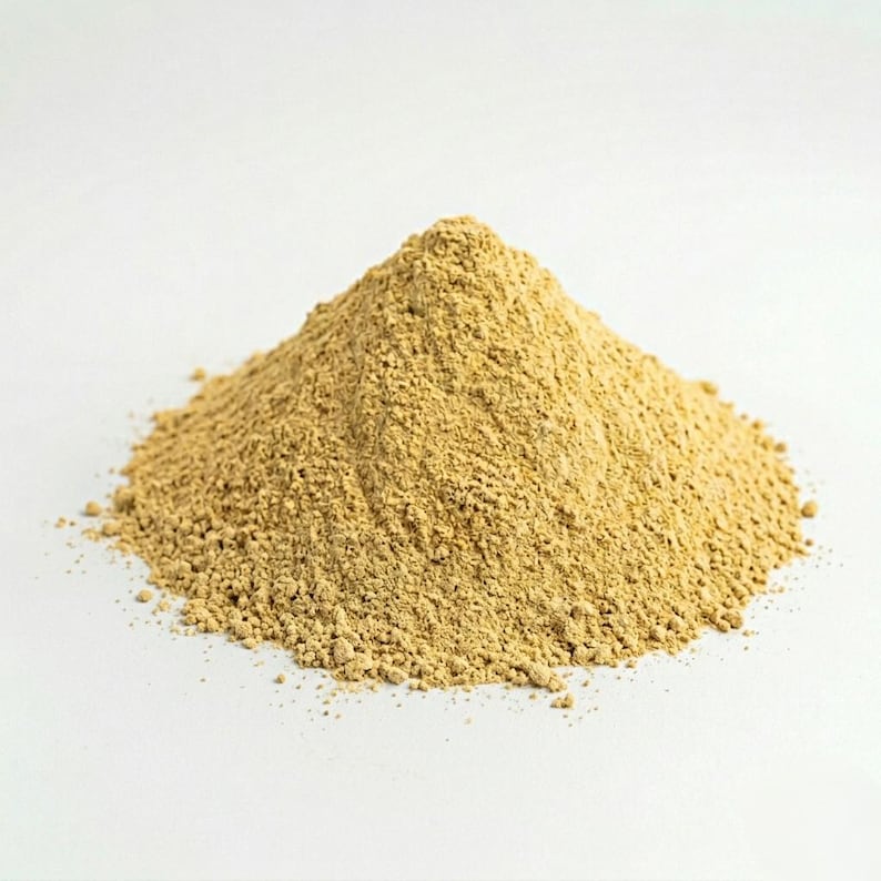 May include: A pile of light brown powder, possibly a spice or ingredient, sits against a white background. The powder is finely ground and has a slightly textured appearance, with a small peak at the top.