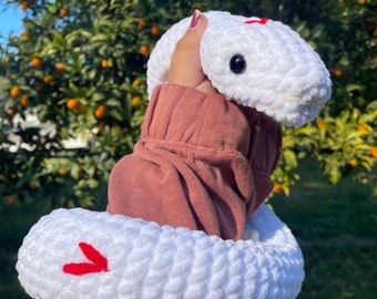 May include: White crocheted snake plush toy with red heart details. The snake is curled up in a person's hand.