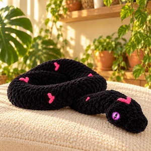 May include: A black crocheted snake plush toy featuring small pink heart-shaped patterns along its body and a single bright purple eye with a vertical slit pupil. The plush is coiled in a circular shape with its head resting on the outer edge.