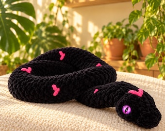 May include: A black crocheted snake plush toy featuring small pink heart-shaped patterns along its body and a single bright purple eye with a vertical slit pupil. The plush is coiled in a circular shape with its head resting on the outer edge.