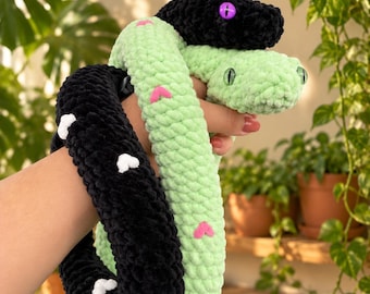 May include: A plush snake toy crafted from soft yarn, featuring two distinct sections: one black with small white heart-shaped accents and one light green with small pink heart-shaped accents. The black head has purple eyes, and the green head has green eyes.