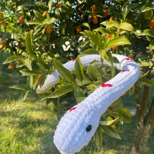 May include: A white crocheted snake with red hearts on its body. The snake is wrapped around a tree branch with green leaves and small orange fruits.