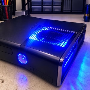 May include: A black Xbox 360 video game console featuring a glossy finish, a central power button illuminated with a bright blue light, and a top ventilation grill glowing with intense blue light, positioned on a light-colored desk surface.