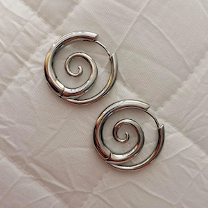 Puede incluir: Un par de pendientes de aro en tono plateado que presentan un diseño de espiral prominente centrado dentro de cada marco circular. Los pendientes tienen un acabado metálico pulido y reflectante con un cierre de bisagra visible.