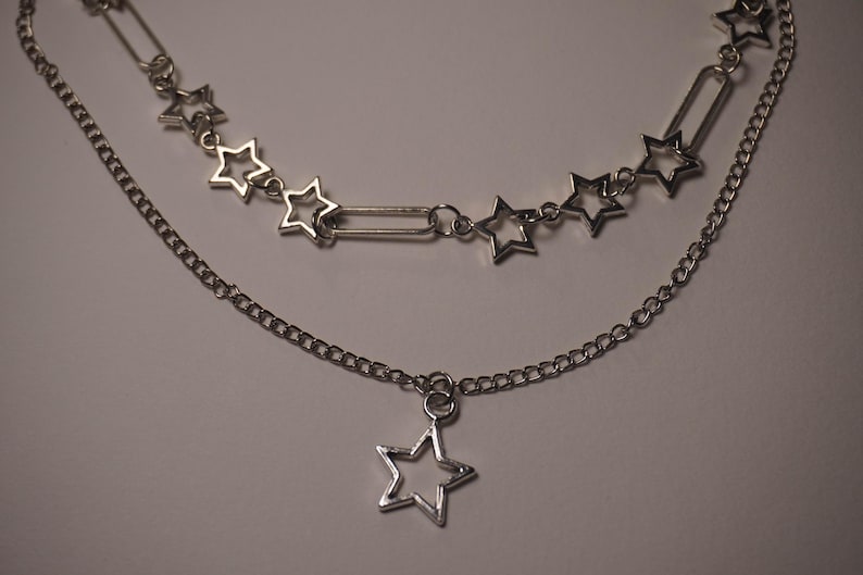 May include: A layered silver-toned necklace featuring two chains. The top chain has star-shaped charms and a rectangular link. The bottom chain has a single star pendant. The jewelry is displayed against a neutral background.