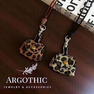 May include: Two small, brown and black leopard print purses with gold accents hang from black and brown cords. The purses are attached to silver clasps. The purses are hanging against a brown background with the text "ARGOTHIC JEWELRY & ACCESSORIES" at the bottom.