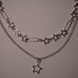 May include: A layered silver-toned necklace with star-shaped pendants. The top layer features star charms connected by safety pin-like links. The bottom layer has a delicate chain with a single star pendant. The necklace is displayed against a neutral background.